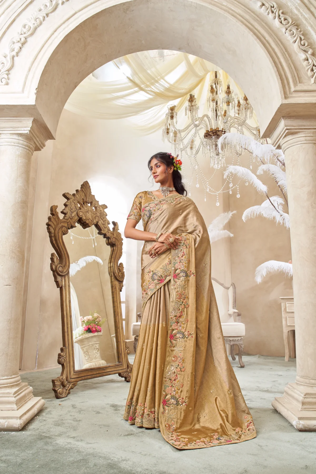 Model posing in a shimmering gold tissue silk designer saree with a heavily embroidered floral border, standing under an ornate archway in a luxurious setting.