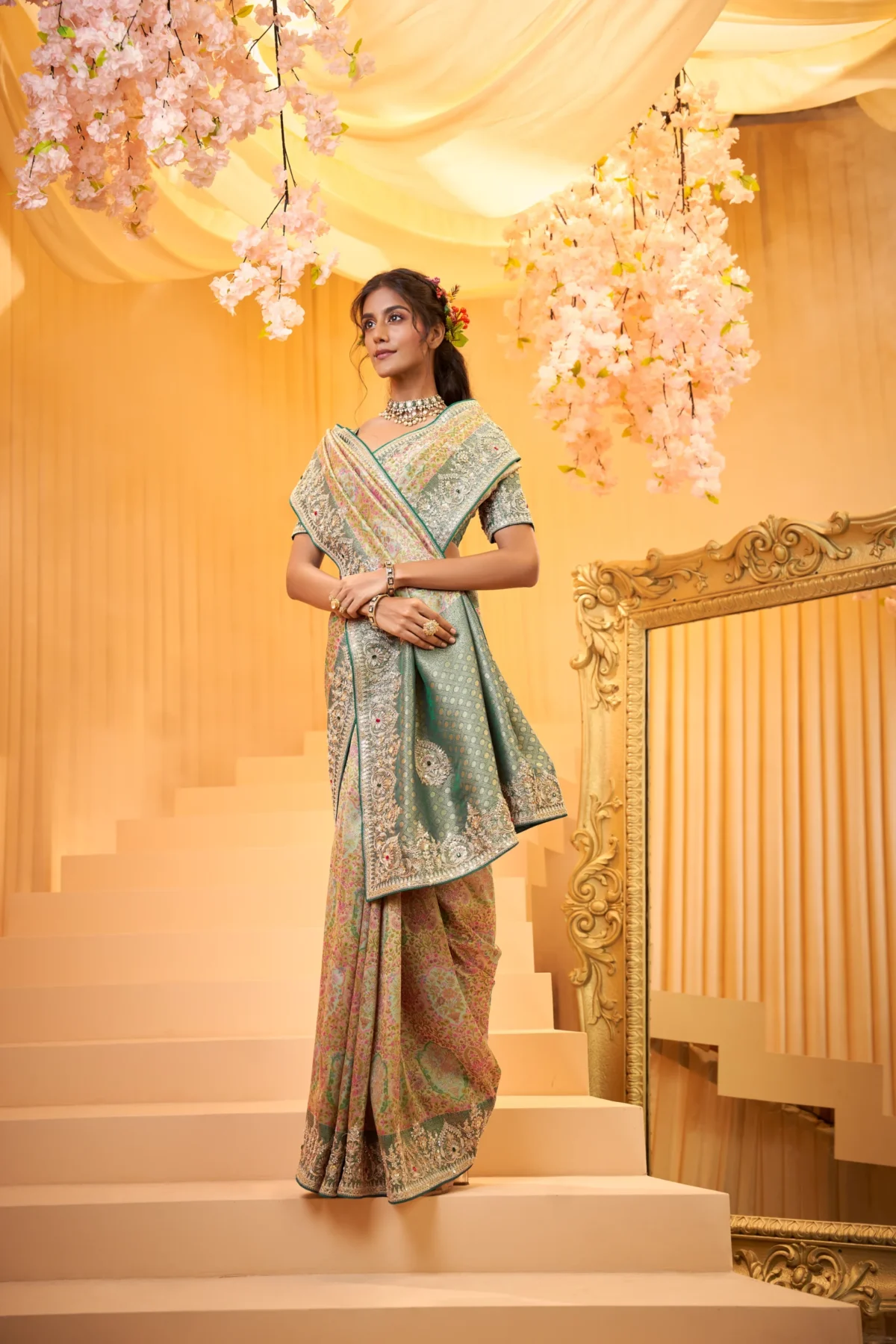 Model posing in a luxurious shimmering gold tissue silk designer saree featuring heavy floral embroidered borders and pallu, standing beneath a grand archway with a chandelier and antique mirror.