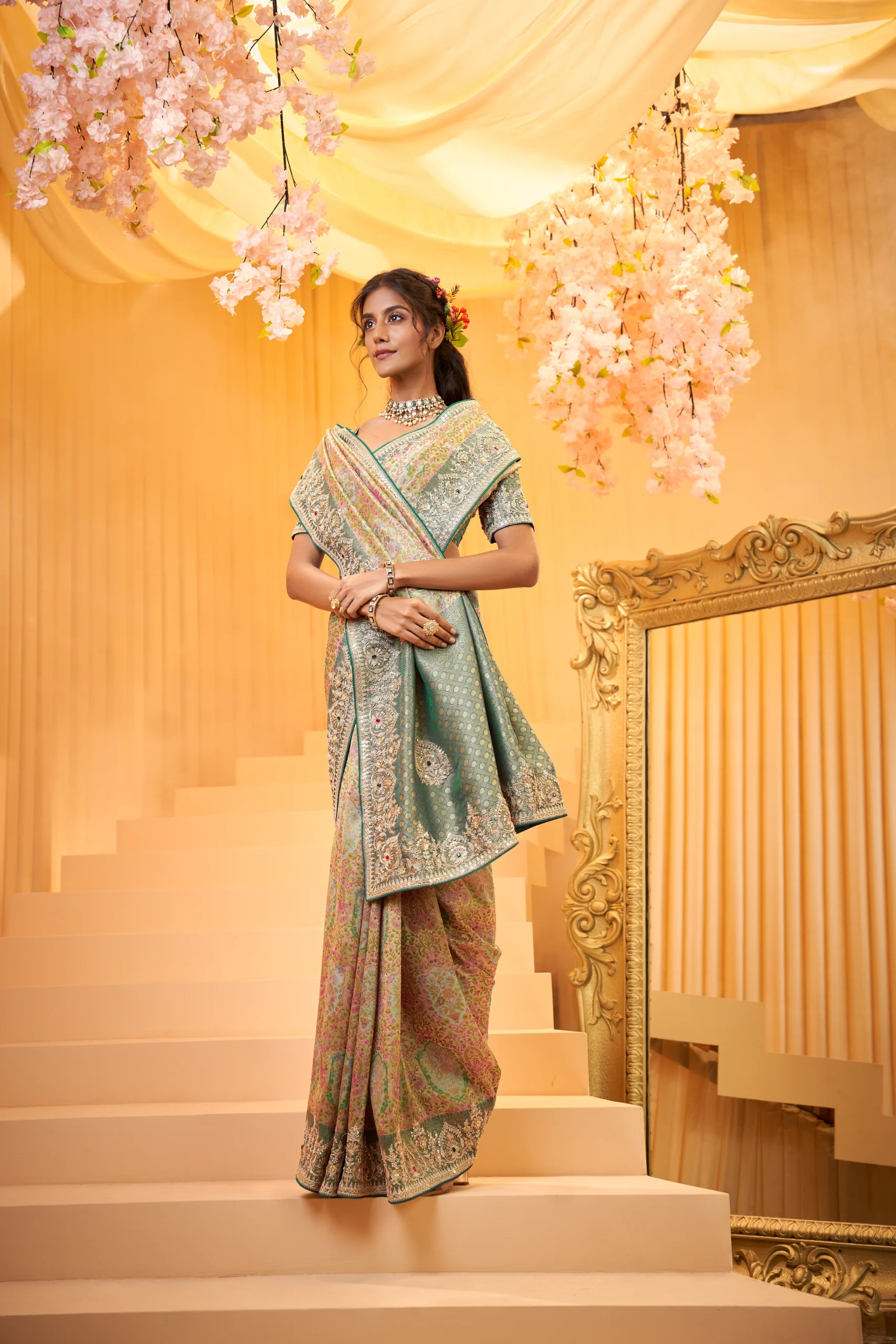 Model posing in a luxurious shimmering gold tissue silk designer saree featuring heavy floral embroidered borders and pallu, standing beneath a grand archway with a chandelier and antique mirror.