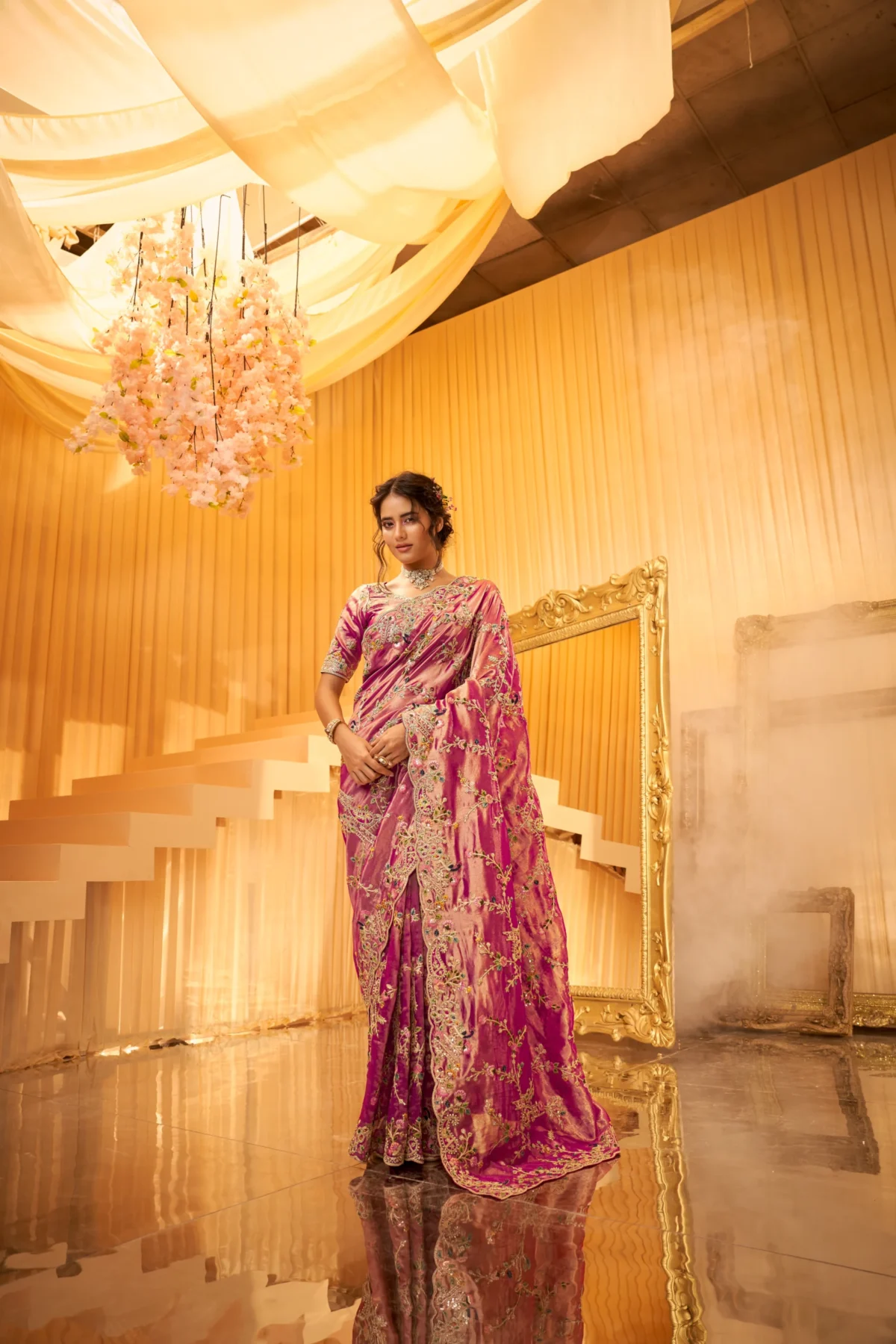 Model posing in a heavy embroidered magenta designer saree with rich gold and silver Zardosi work in a luxurious golden setting.