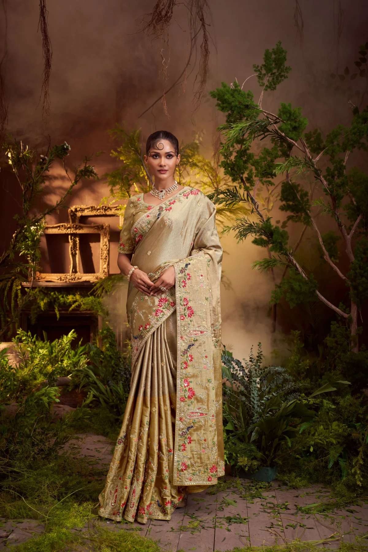 Model posing in an antique gold designer saree with multi-color floral and sequin embroidery, framed by a large ornate golden frame.
