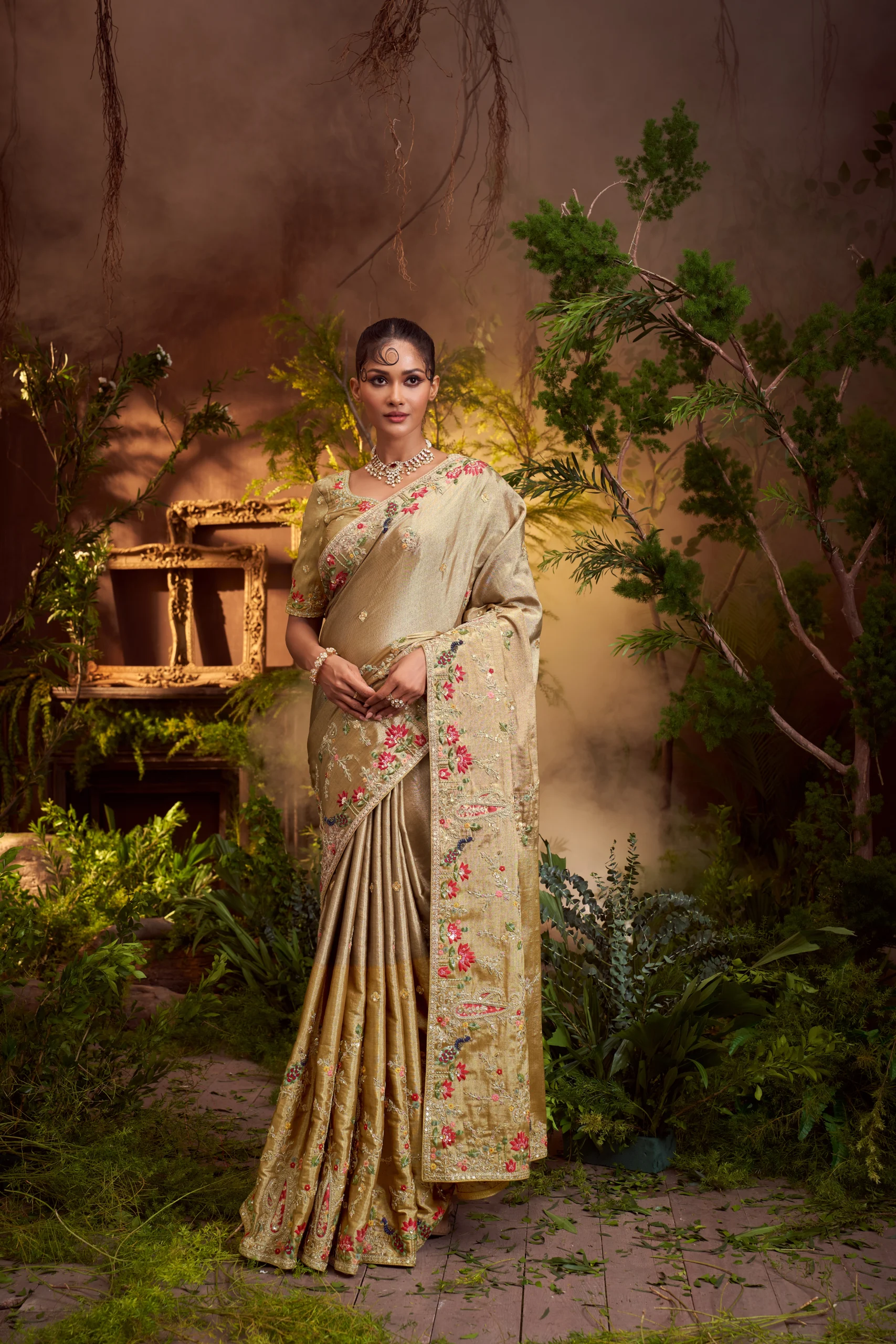 Model posing in an antique gold designer saree with multi-color floral and sequin embroidery, framed by a large ornate golden frame.