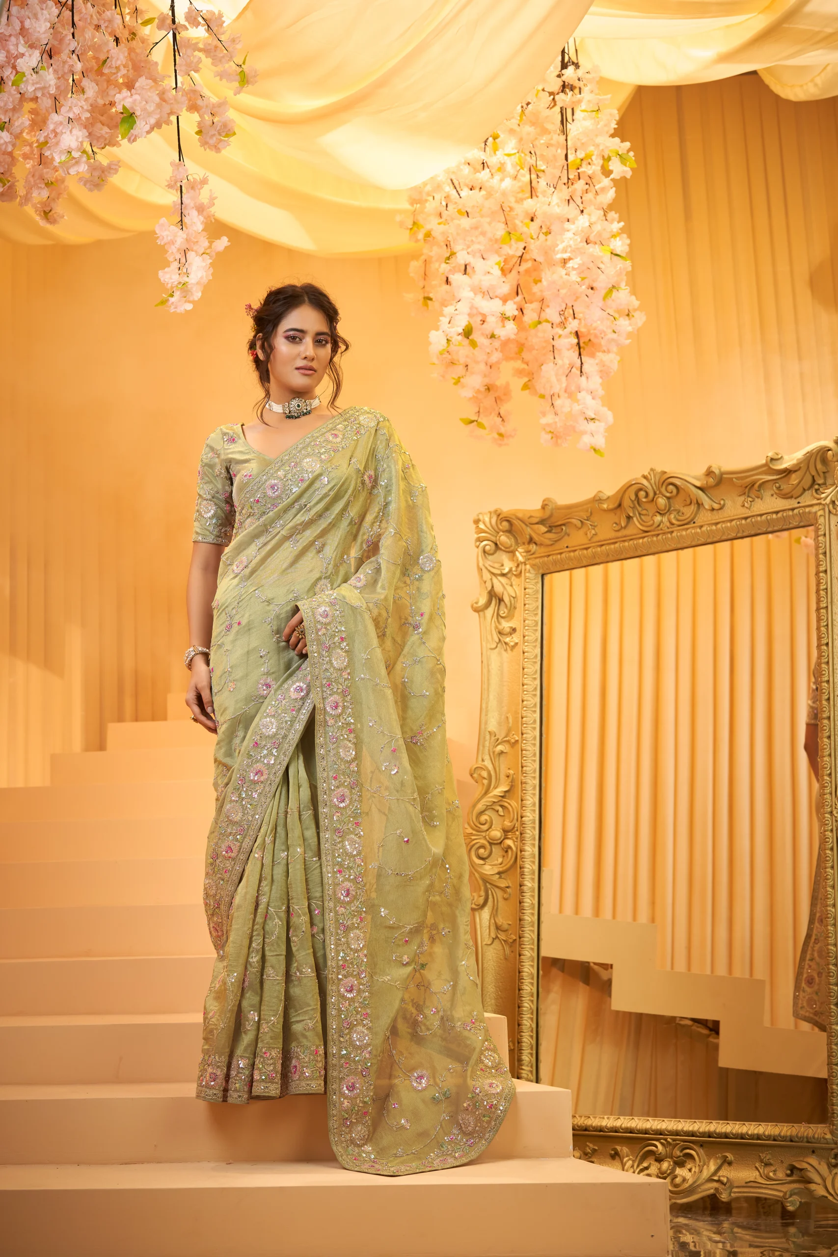 Model posing on a staircase wearing a luxurious lime green designer silk saree featuring heavy floral embroidery and embellished gold borders for a wedding function.