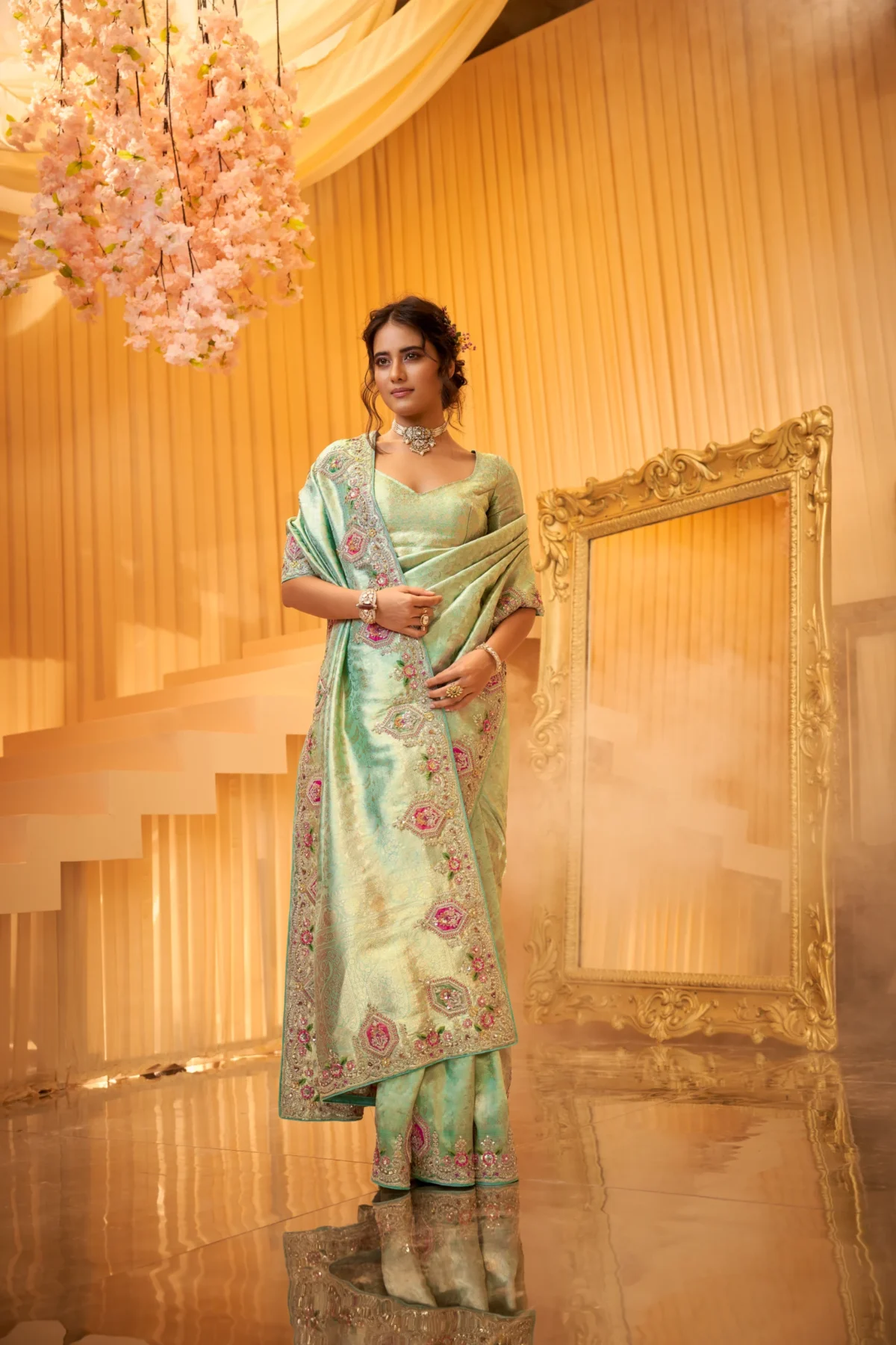 A model posing in a heavily embroidered light jade green designer silk saree with pink floral details and silver zari work in a golden-hued luxury setting.